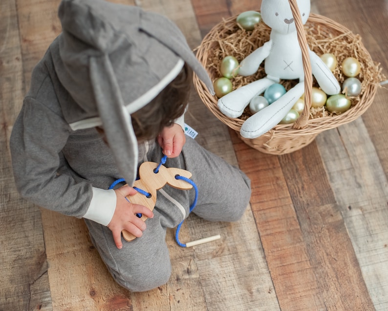 May include: A child wearing a gray bunny hooded onesie plays with a wooden bunny lacing toy. The toy has a blue string threaded through the holes. The child is sitting on a wooden floor.