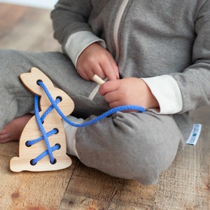 May include: A wooden bunny toy with blue laces for threading. The toy is on a wooden floor and a child is playing with it.