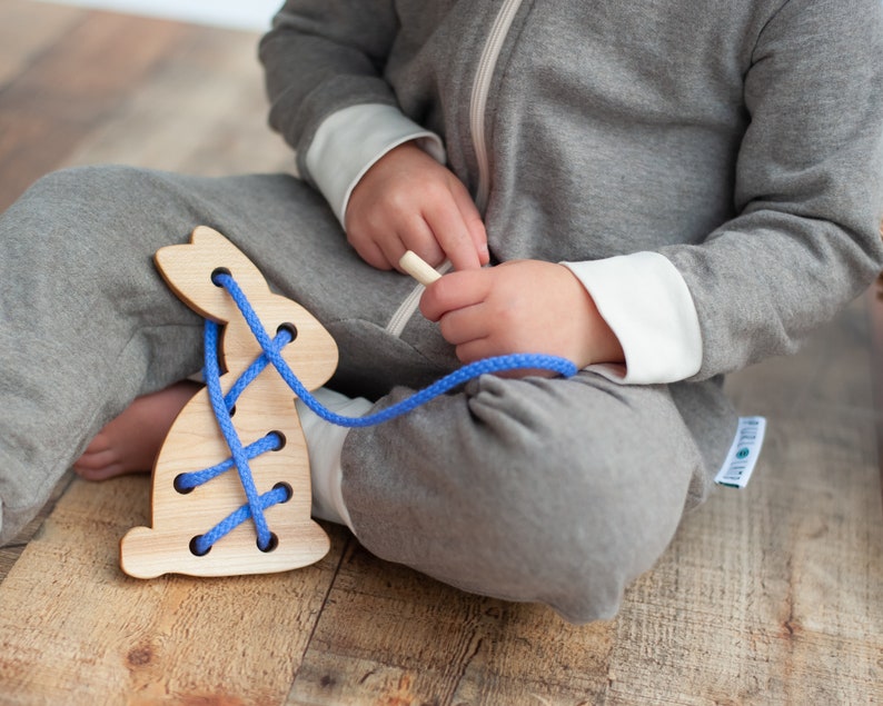 May include: A wooden bunny toy with blue laces for threading. The toy is on a wooden floor and a child is playing with it.