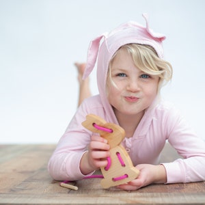 May include: A young girl wearing a pink bunny hooded sweatshirt plays with a wooden bunny lacing toy. The toy has pink string threaded through the holes.