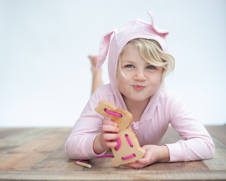 May include: A young girl wearing a pink bunny hooded sweatshirt plays with a wooden bunny lacing toy. The toy has pink string threaded through the holes.