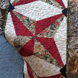 May include: A quilted table runner with a starburst design. The runner features red, cream, and green fabrics with a floral pattern. The edges are finished with a green and cream border.
