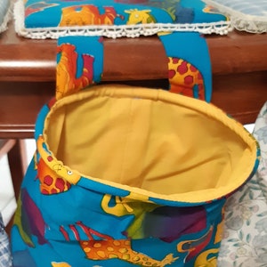 May include: A fabric storage bag with a bright blue exterior featuring a giraffe design. The bag has a yellow lining and blue handles. A yellow pincushion and a matching fabric square with white lace trim are also visible.