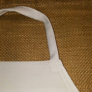 May include: Close-up of a white apron with a brown burlap background. The apron has a white strap and is made of a soft, woven fabric.