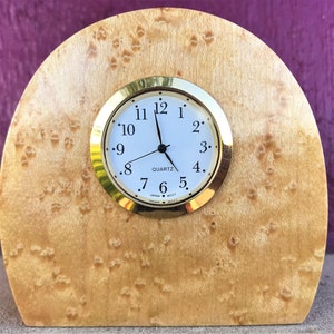 Op de afbeelding: Een houten klok met een gouden rand om de wijzerplaat en zwarte wijzers. De klok staat op een houten voet met een burl-houtpatroon.