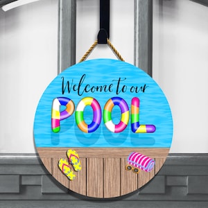 May include: Round wooden door hanger with a blue pool background and the text "Welcome to our POOL" in black lettering. The word "POOL" is made up of colorful inflatable pool rings. The bottom of the hanger shows a wooden deck with flip flops and a towel.