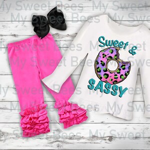 Puede incluir: Una camiseta blanca de manga larga con un gráfico de donut rosa y verde y el texto "Sweet & Sassy". La camiseta se combina con leggings rosas con volantes en la parte inferior y un lazo negro.