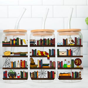 May include: Three clear glass drinking cups with bamboo lids and glass straws. Each cup features a bookshelf design with various themed illustrations, including a broom, owl, and sorting hat. The cups are on a white tiled surface.