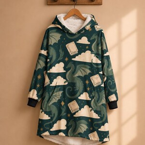 May include: A dark green hooded sweatshirt featuring a repeating pattern of light green dragons, cream-coloured clouds, books, and small stars. The garment has a white sherpa fleece lining, a hood, long sleeves with black cuffs, and a high-low hemline.