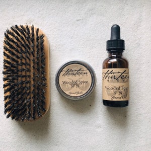 May include: A set of beard care products in a woodsy scent. The set includes a wooden brush, a small tin of beard balm, and a glass bottle of beard oil. All three products feature the label "Thirteen Thieves" and the scent "Wooded Spice No. 29".