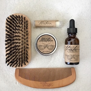 Może przedstawiać: Zestaw produktów do pielęgnacji brody w aromacie Tobacco Bourbon. Zestaw zawiera drewniany grzebień, szczotkę, puszkę balsamu do brody i butelkę olejku do brody. Wszystkie produkty mają logo "Thirteen Thieves".
