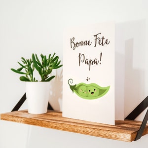 May include: A white card with the text 'Bonne Fete Papa!' and a cartoon image of two green peas with faces in a pea pod. The card is sitting on a wooden shelf with a small potted succulent plant.