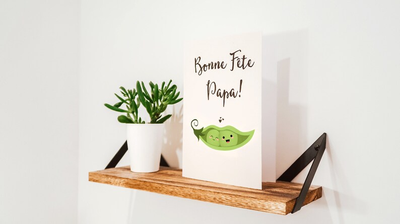 May include: A white card with the text 'Bonne Fete Papa!' and a cartoon image of two green peas with faces in a pea pod. The card is sitting on a wooden shelf with a small potted succulent plant.