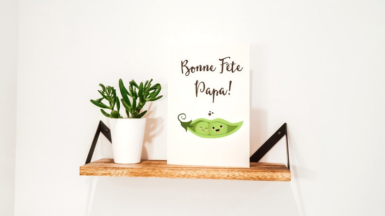 May include: A white card with the text 'Bonne Fete Papa!' in black script.  Two green peas with faces are inside a green pea pod. The card is sitting on a wooden shelf with a small green succulent plant in a white pot.