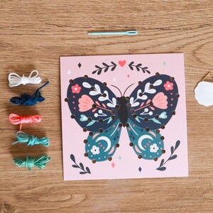 May include: A pink card with a butterfly design. The butterfly has blue, green, and pink floral patterns. The card has a white circle with a gold hanger. There are five spools of thread in white, blue, orange, green, and teal. There is a white strip with 15 white circles and a blue needle.