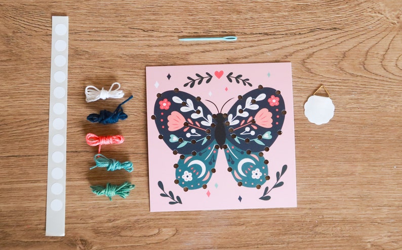 May include: A pink card with a butterfly design. The butterfly has blue, green, and pink floral patterns. The card has a white circle with a gold hanger. There are five spools of thread in white, blue, orange, green, and teal. There is a white strip with 15 white circles and a blue needle.