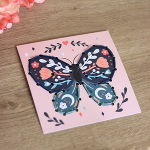 May include: A pink card with a blue butterfly with floral details. The butterfly is outlined with black stitching. The card has a heart and floral designs in the background.