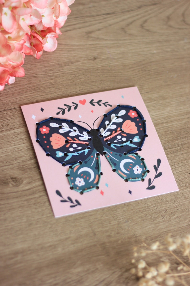 May include: A pink card with a blue butterfly with floral details. The butterfly is outlined with black stitching. The card has a heart and floral designs in the background.