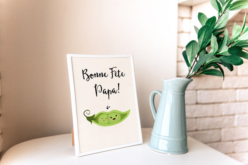 May include: A white picture frame with a white mat and a white background. The frame is standing on a white surface. The image in the frame is a cartoon of two peas in a pea pod. The peas are smiling and have eyes. The text on the image reads 'Bonne Fete Papa!'.