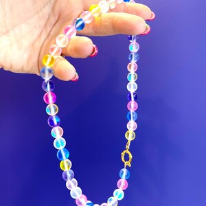 Op de afbeelding: Een kleurrijke kralenketting met doorschijnende, iriserende kralen in roze, geel, blauw en paars. De ketting wordt getoond tegen een blauwe achtergrond, met een goudkleurige sluiting.