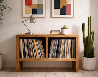 May include: A wooden record cabinet filled with vinyl records, flanked by a tall cactus in a white pot and a potted tree. Two framed abstract art prints hang above the cabinet, and a silver desk lamp sits on top.