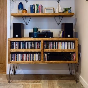 May include: A wooden media console with hairpin legs, filled with CDs. Above, two shelves display books, decorative items, and plants. The shelves are supported by black metal brackets, creating a stylish storage solution.