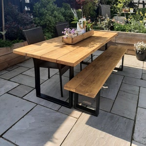 May include: A wooden outdoor dining table and bench set with black metal legs. The table is set with a wooden planter box filled with flowers and greenery. The table and bench are made of natural wood with a light brown finish.