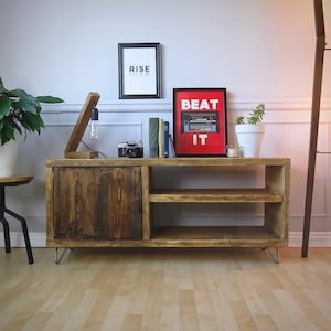 K&ouml;nnte beinhalten: Rustikaler TV-Schrank aus Holz mit T&uuml;r und offenen Regalen. Ein gerahmter Druck mit dem Text "BEAT IT" h&auml;ngt dar&uuml;ber. Eine Holzlampe mit Gl&uuml;hbirne und eine Vintage-Kamera stehen darauf. Eine Stehlampe und Topfpflanzen sind ebenfalls im Raum. Der Schrank ist etwa 120 cm breit.
