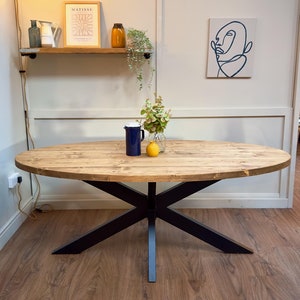 May include: Oval wooden dining table with a natural finish, supported by a black metal base. The table is decorated with a blue jug, a vase of flowers, and a lemon. The table is approximately 183 cm long.