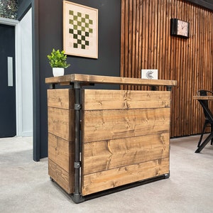 May include: A wooden reception desk with a light brown finish. The desk has a rectangular design with metal supports and a top surface. The wood grain is visible, and the desk is set against a dark wall with a framed artwork.