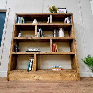 Puede incluir: Una estanter&iacute;a grande de madera r&uacute;stica con m&uacute;ltiples compartimentos, llena de libros, objetos decorativos y una m&aacute;quina de escribir vintage. La estanter&iacute;a est&aacute; te&ntilde;ida de un marr&oacute;n c&aacute;lido y se apoya ligeramente en la pared. La habitaci&oacute;n tiene una paleta de colores neutros.