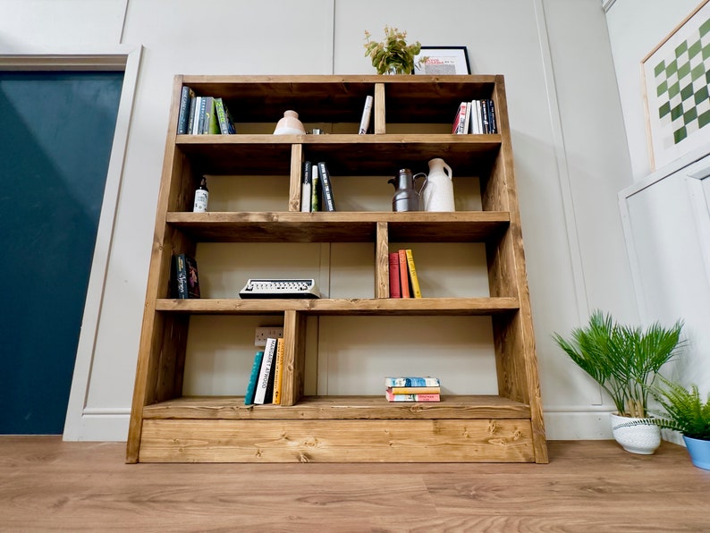 Puede incluir: Una estanter&iacute;a grande de madera r&uacute;stica con m&uacute;ltiples compartimentos, llena de libros, objetos decorativos y una m&aacute;quina de escribir vintage. La estanter&iacute;a est&aacute; te&ntilde;ida de un marr&oacute;n c&aacute;lido y se apoya ligeramente en la pared. La habitaci&oacute;n tiene una paleta de colores neutros.