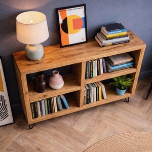 Op de afbeelding: Een houten boekenkast met meerdere compartimenten, gevuld met boeken en decoratieve items. Bovenop staat een lamp met een witte kap en een ingelijste abstracte kunstprint is zichtbaar. De boekenkast wordt ondersteund door zwarte metalen poten.