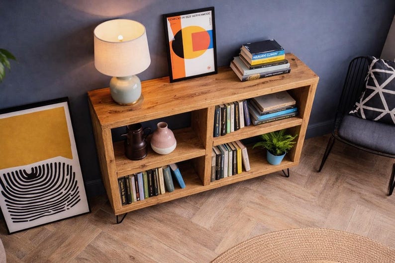 Op de afbeelding: Een houten boekenkast met meerdere compartimenten, gevuld met boeken en decoratieve items. Bovenop staat een lamp met een witte kap en een ingelijste abstracte kunstprint is zichtbaar. De boekenkast wordt ondersteund door zwarte metalen poten.