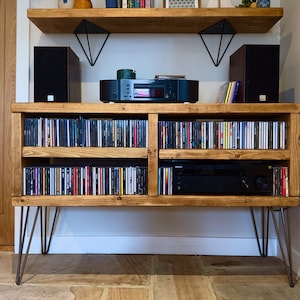 May include: Wooden media console with hairpin legs, filled with CDs. The console has two shelves for storage and a top shelf with speakers and audio equipment. The wood has a natural finish, and the legs are metal.
