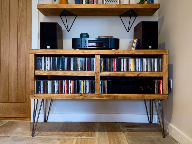 May include: Wooden media console with hairpin legs, filled with CDs. The console has two shelves for storage and a top shelf with speakers and audio equipment. The wood has a natural finish, and the legs are metal.