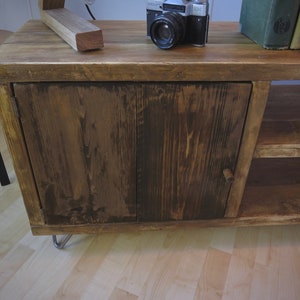 K&ouml;nnte beinhalten: Ein rustikaler Holzschrank mit dunkler Beize. Der Schrank hat eine einzelne T&uuml;r mit einem Holzknauf und ein unteres Regal. Eine Vintage-Kamera und B&uuml;cher liegen obenauf. Der Schrank wird von Metall-Haarnadelbeinen getragen.