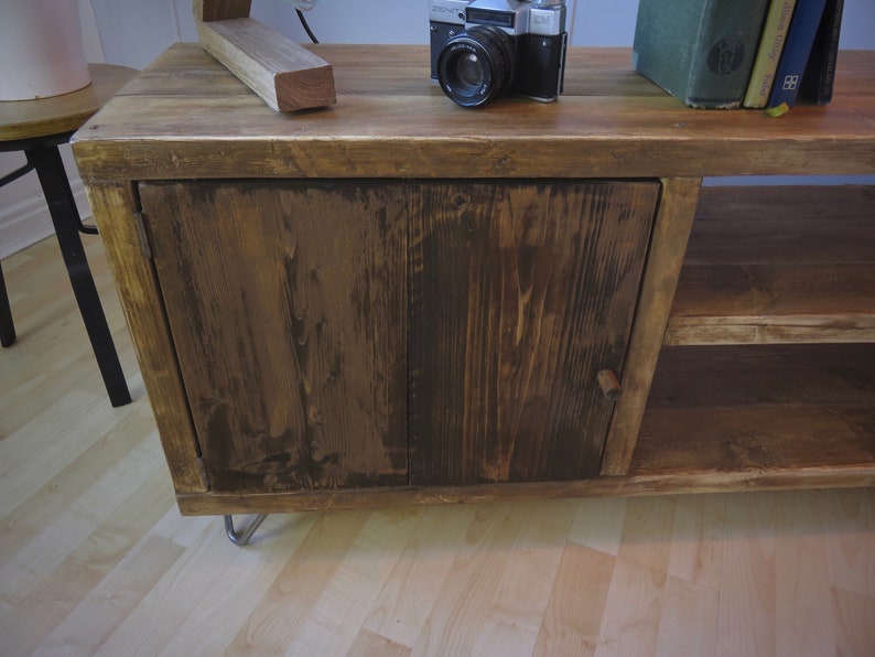 K&ouml;nnte beinhalten: Ein rustikaler Holzschrank mit dunkler Beize. Der Schrank hat eine einzelne T&uuml;r mit einem Holzknauf und ein unteres Regal. Eine Vintage-Kamera und B&uuml;cher liegen obenauf. Der Schrank wird von Metall-Haarnadelbeinen getragen.