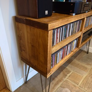 May include: A rustic, wooden media console with a warm, honey-toned finish. The console features two shelves filled with CDs and is supported by metal hairpin legs. A speaker and audio equipment sit on top.