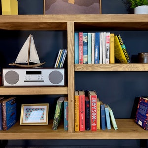 May include: Wooden bookshelf with various books, a small sailboat model, a framed picture, and a stereo. The books have colorful spines and titles. Decorative items include a blue vase and a small orange jar. The shelf is made of light-colored wood.