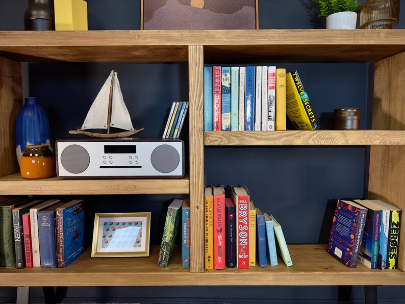 May include: Wooden bookshelf with various books, a small sailboat model, a framed picture, and a stereo. The books have colorful spines and titles. Decorative items include a blue vase and a small orange jar. The shelf is made of light-colored wood.