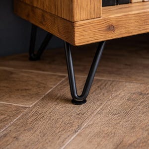 Op de afbeelding: Close-up van een houten kast met zichtbare houtnerf. De kast heeft zwarte metalen haarspeldpoten. De poten zijn bevestigd aan de onderkant van de kast. De kast staat op een houten vloer.