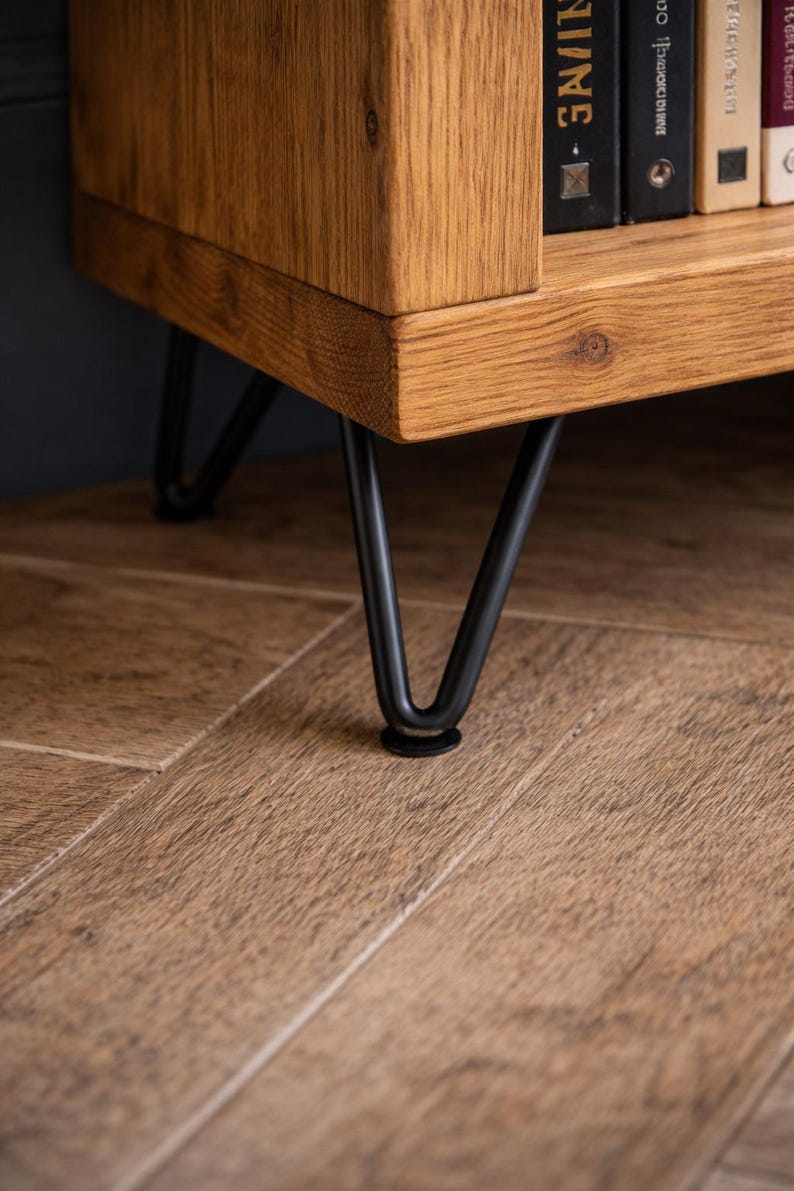 Op de afbeelding: Close-up van een houten kast met zichtbare houtnerf. De kast heeft zwarte metalen haarspeldpoten. De poten zijn bevestigd aan de onderkant van de kast. De kast staat op een houten vloer.