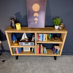 May include: A wooden bookshelf with a variety of items. A framed print of the moon phases hangs above the shelf. The shelf displays books, decorative vases, a small sailboat, a radio, and a small potted plant.