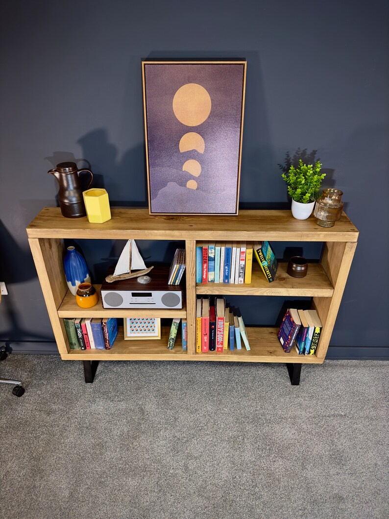 May include: A wooden bookshelf with a variety of items. A framed print of the moon phases hangs above the shelf. The shelf displays books, decorative vases, a small sailboat, a radio, and a small potted plant.