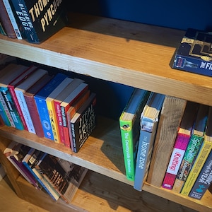 May include: A wooden bookshelf with multiple shelves filled with various books. The books are organized by color and genre. The top shelf has a book titled "The First Fifteen Lives of Harry August".