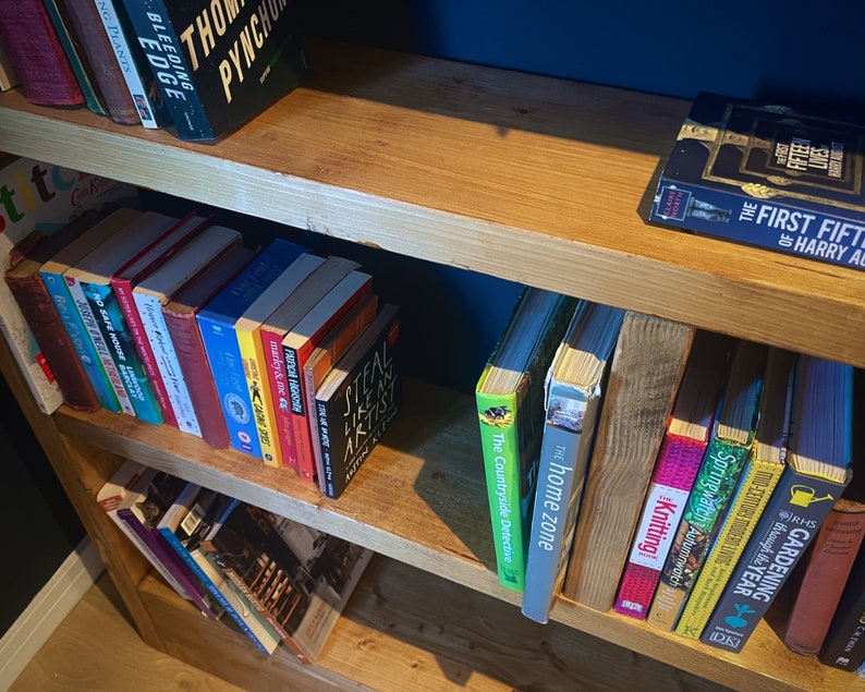 May include: A wooden bookshelf with multiple shelves filled with various books. The books are organized by color and genre. The top shelf has a book titled "The First Fifteen Lives of Harry August".
