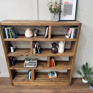 Puede incluir: Estanter&iacute;a de madera marr&oacute;n con m&uacute;ltiples estantes, llena de libros, objetos decorativos y una m&aacute;quina de escribir vintage. La estanter&iacute;a tiene un dise&ntilde;o asim&eacute;trico. Una impresi&oacute;n enmarcada y un jarr&oacute;n con vegetaci&oacute;n est&aacute;n encima.