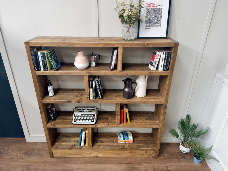 Puede incluir: Estanter&iacute;a de madera marr&oacute;n con m&uacute;ltiples estantes, llena de libros, objetos decorativos y una m&aacute;quina de escribir vintage. La estanter&iacute;a tiene un dise&ntilde;o asim&eacute;trico. Una impresi&oacute;n enmarcada y un jarr&oacute;n con vegetaci&oacute;n est&aacute;n encima.