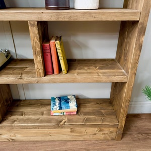 Puede incluir: Estanter&iacute;a r&uacute;stica de madera con tres estantes. Los estantes exhiben libros de varios colores, una m&aacute;quina de escribir vintage y objetos decorativos. La madera tiene un tono marr&oacute;n c&aacute;lido, con un dise&ntilde;o sencillo y funcional.